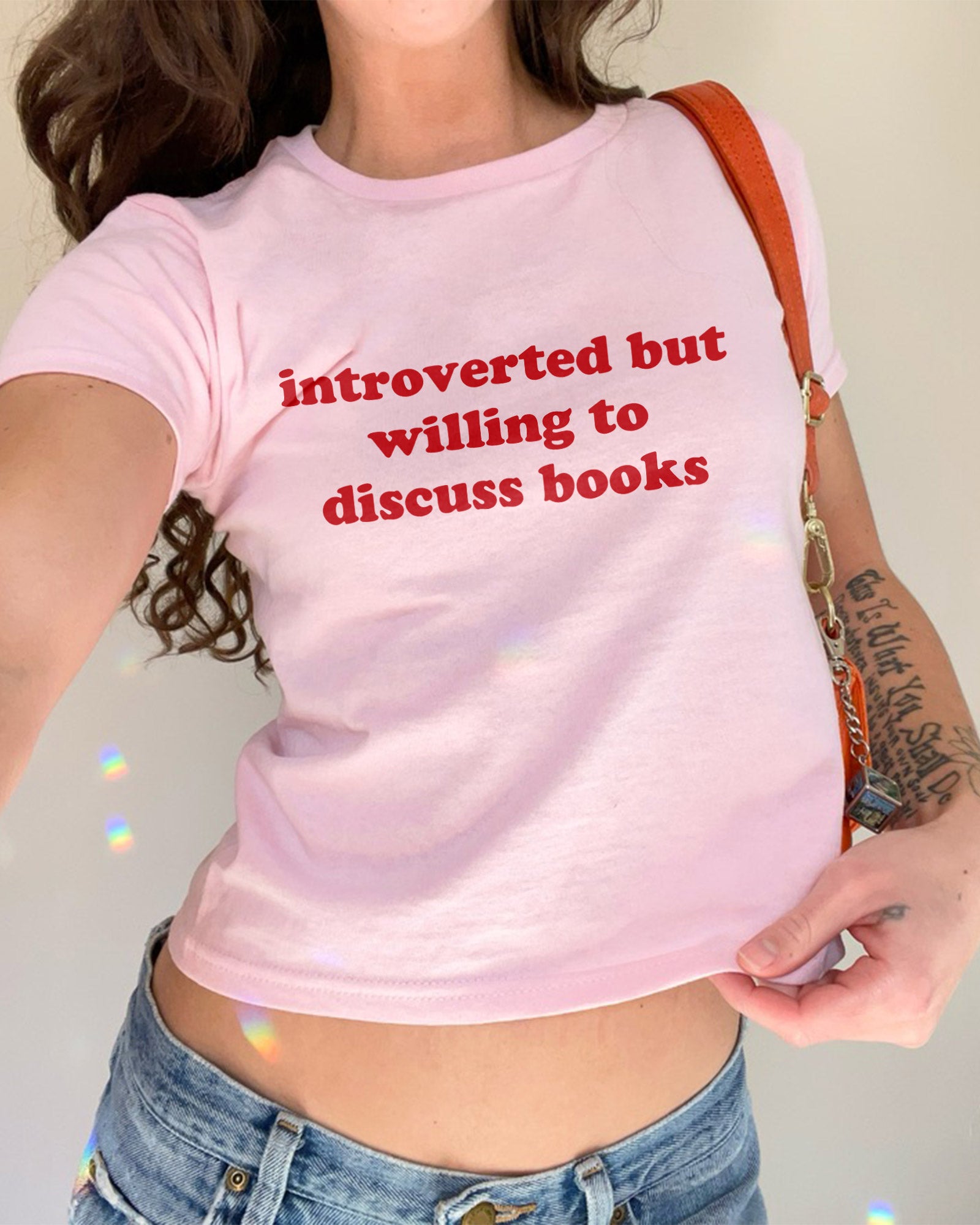 Woman wearing light pink baby tee with “Introverted But Willing To Discuss Books” quote